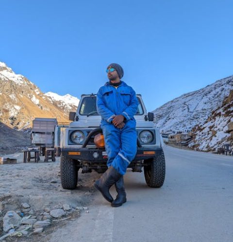 A man in a blue suit poses confidently by an SUV on a snowy mountain road in Manali.