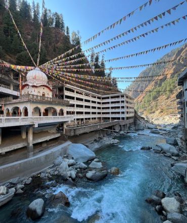Colorful flags draped over Manikaran temple by a rushing river in Himachal Pradesh, India.